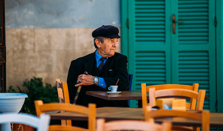 An older man wearing a hat sits in an outdoor cafe enjoying a coffee