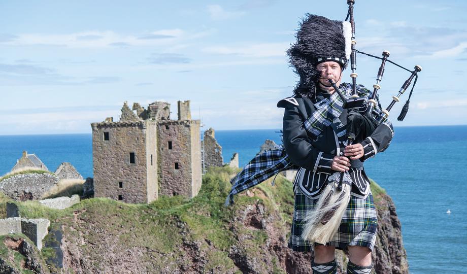 Scottish piper stands next to a castle overlooking the ocean