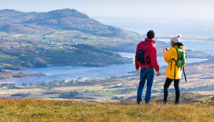 Two hikers overlooking a scenic Irish landscape with mountains and a shimmering lake in the distance.